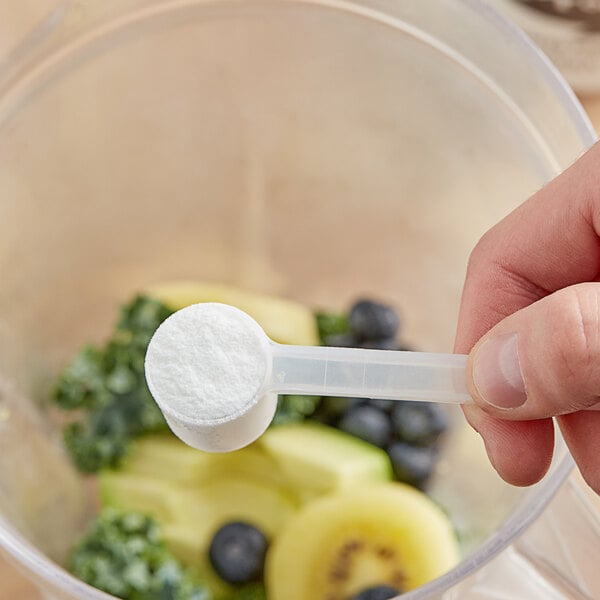 A hand holding a spoon with a white powder over a blender.