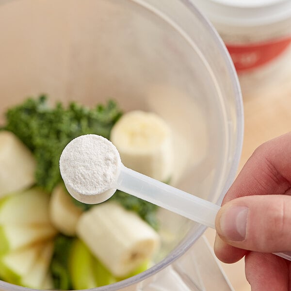 A person using a spoon to add Add A Scoop Energy Blend powder to a blender.