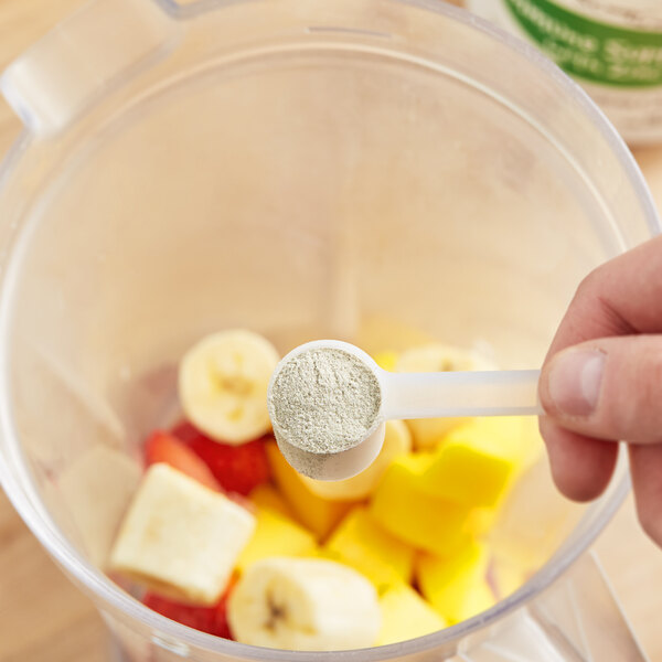 A hand holding a spoon with Add A Scoop Immune Support Blend supplement powder in a blender with fruit.