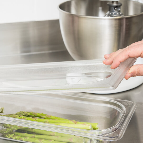 A hand using a Cambro clear polycarbonate flat lid on a plastic food container filled with asparagus.
