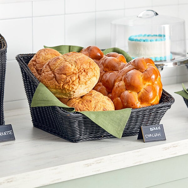 An Acopa black woven plastic rattan basket filled with bread and rolls on a counter.