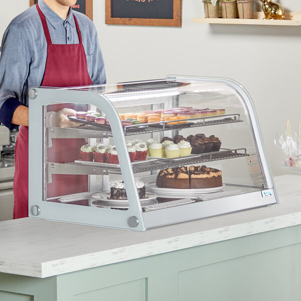 A man in an apron standing behind a white Avantco countertop bakery display case filled with cupcakes and cakes.