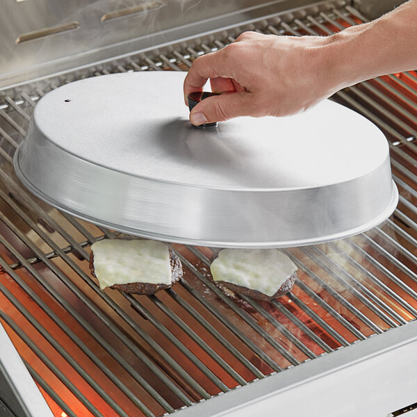 A person using an American Metalcraft aluminum basting cover to cook hamburger patties on a grill.