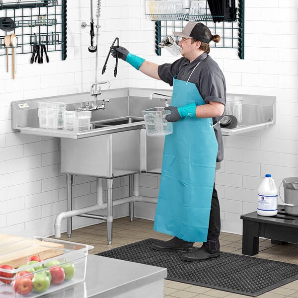 A man in a blue apron and gloves standing at a Regency stainless steel 3 compartment sink.