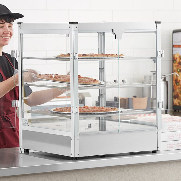 A woman wearing a black apron and hat using a Carnival King countertop warmer to display pizzas.