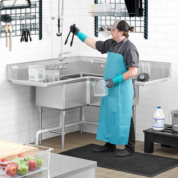 A man in a blue apron using a Regency stainless steel 3 compartment sink in a professional kitchen.