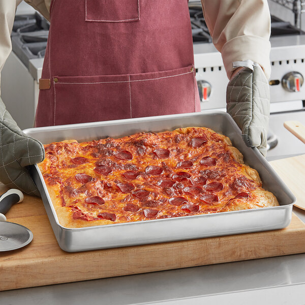 A person in an apron holding a rectangular pizza in an American Metalcraft perforated square pizza pan.