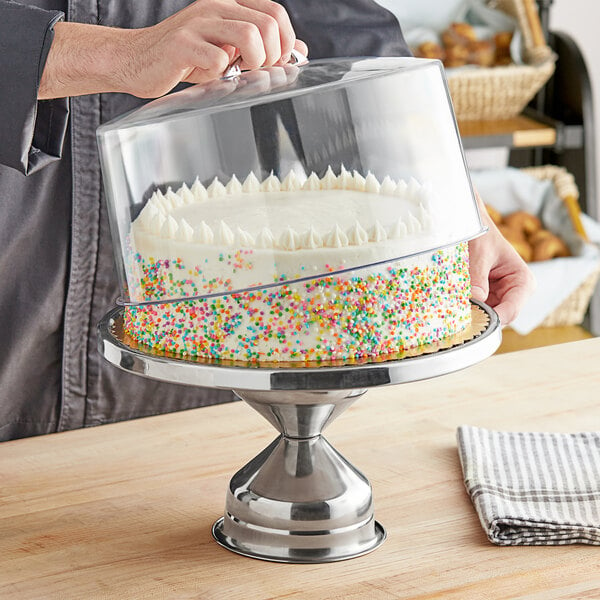 A person holding a cake on a stainless steel cake stand with a clear round cover.