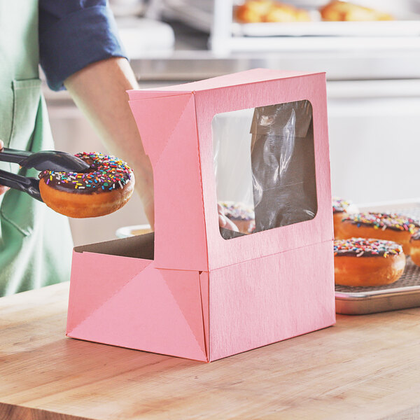 A person holding a chocolate doughnut with sprinkles in a pink bakery box with a window.