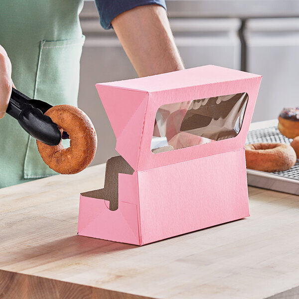 A person using black tongs to place a donut in a Baker's Mark pink window box.
