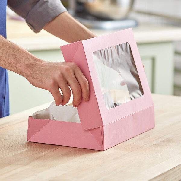 A person opening a Baker's Mark pink bakery box with a clear plastic window on a counter.
