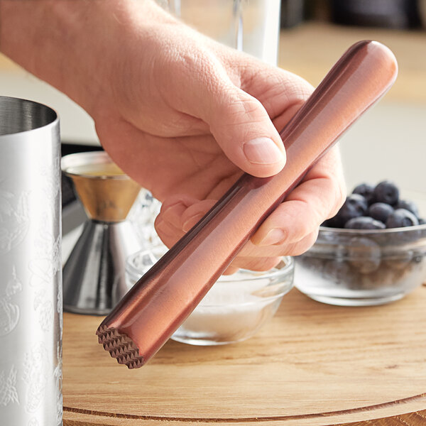 A person using an Acopa copper stainless steel muddler with a tenderizer head to mix blueberries in a glass.