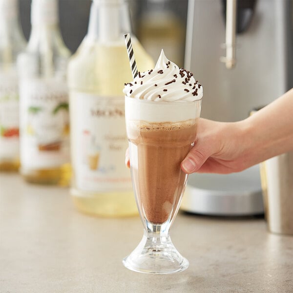 A hand holding a glass of Monin chocolate milkshake with whipped cream and chocolate chips.