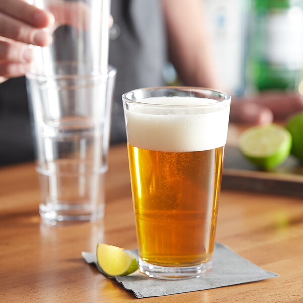 A person holding a clear Acopa Select Stackable Cooler / Mixing Glass filled with beer with foam and a lime wedge.