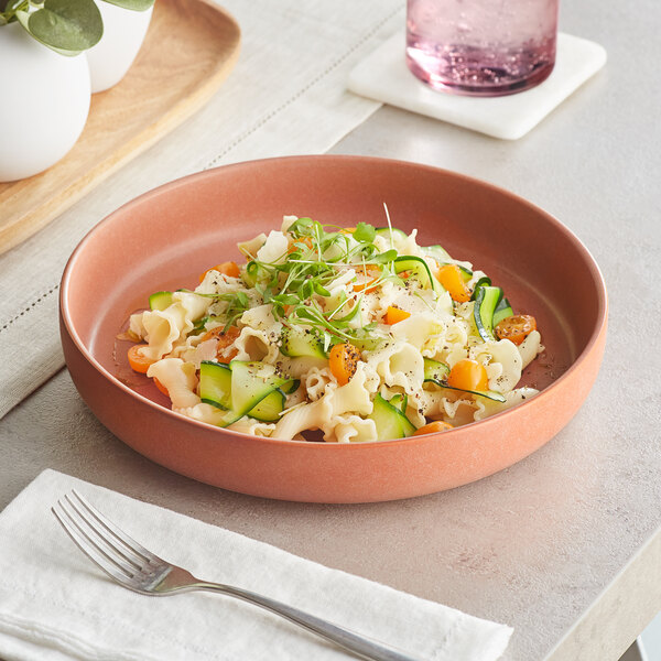 An Acopa Terra Cotta matte porcelain pasta bowl filled with pasta, vegetables, and greens on a table.