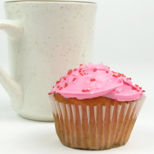 A cupcake with pink frosting and sprinkles in a white fluted baking cup next to a white mug.