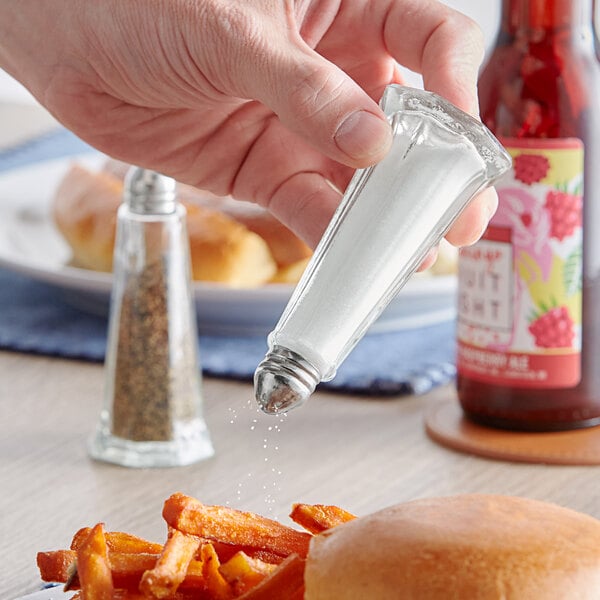 A hand holding a Choice silver Eiffel Tower salt shaker over a plate of fries.