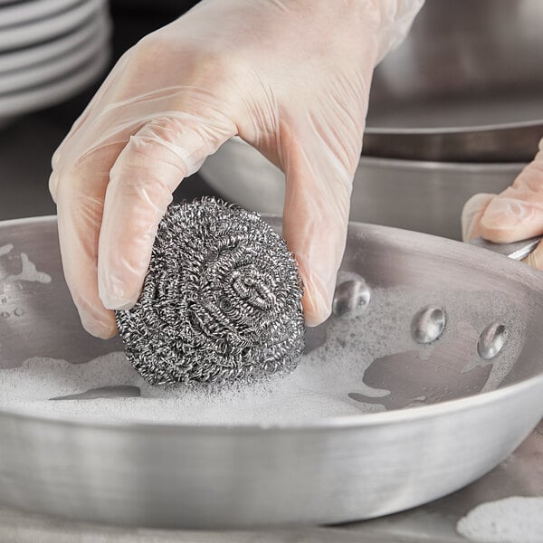 A gloved hand uses a Choice stainless steel scrubber to clean a metal pan.