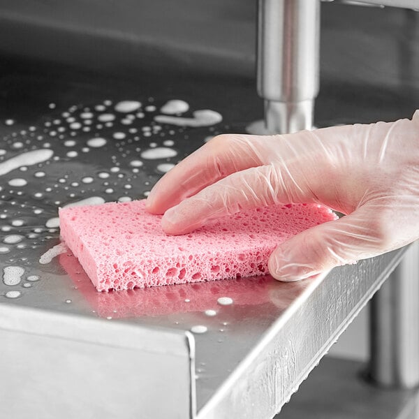 A hand in a glove cleaning a kitchen sink with a pink Lavex cellulose sponge.
