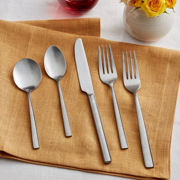 A Acopa Petra stainless steel flatware set on a napkin with yellow roses on a table.