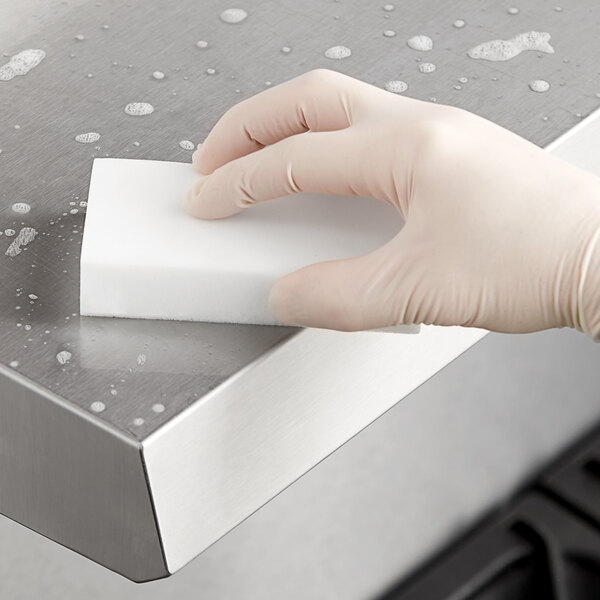 A gloved hand using a white Lavex eraser sponge to clean a stainless steel counter.
