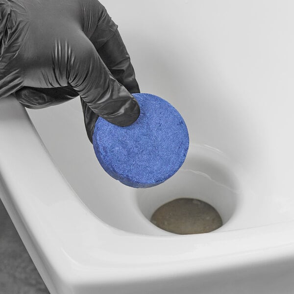 A hand in a black glove holding a blue round Lavex urinal cake over a toilet.