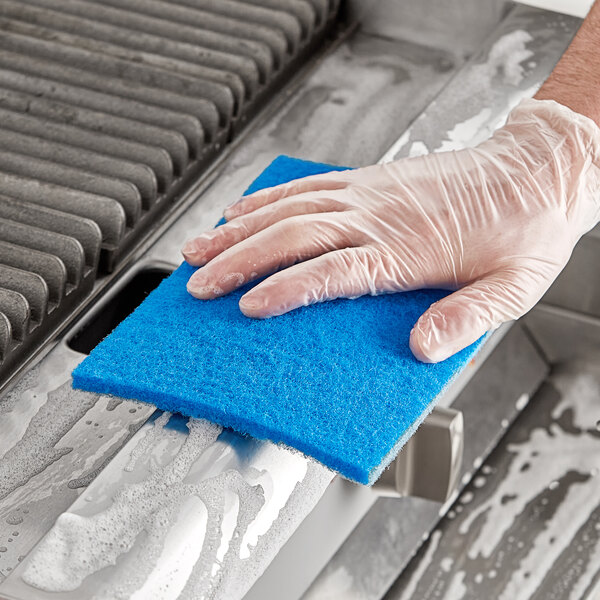 A person wearing gloves cleaning a grill with a blue Lavex light-duty scouring pad.