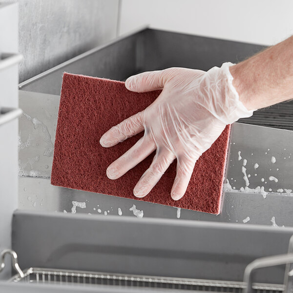 A gloved hand uses a Lavex maroon scouring pad to clean a kitchen counter.
