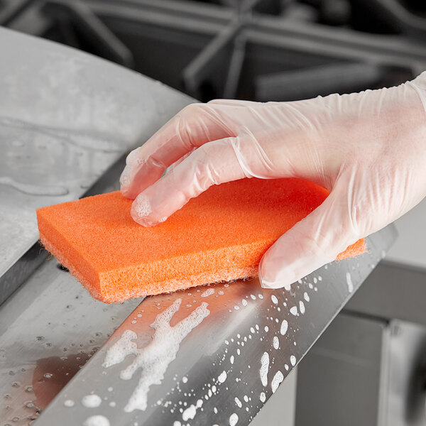 A hand in a rubber glove using an orange Lavex sponge to clean a stainless steel sink.