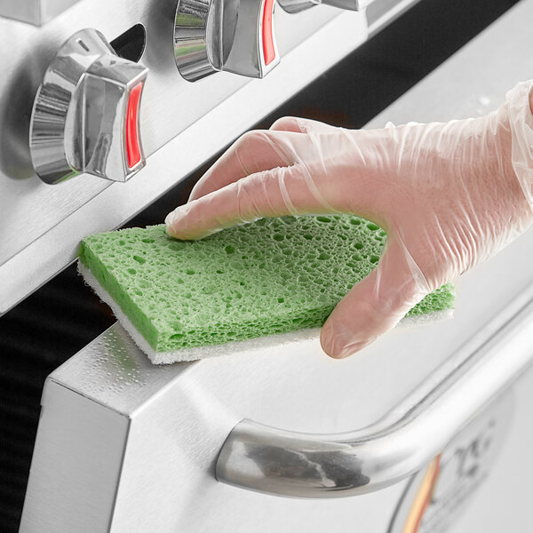 A hand in a glove holding a green Lavex sponge over a stove.