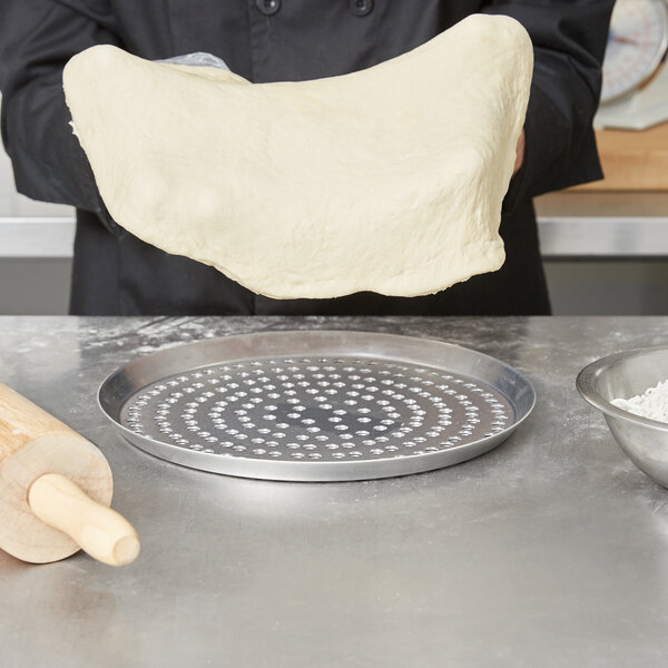 A person holding a piece of dough over an American Metalcraft super perforated pizza pan with a rolling pin.