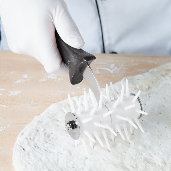 A gloved hand uses an American Metalcraft dough docker with black plastic pins to pierce pizza dough.