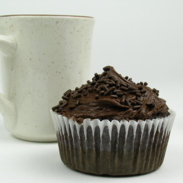 A white fluted baking cup with a chocolate cupcake with chocolate frosting and sprinkles.