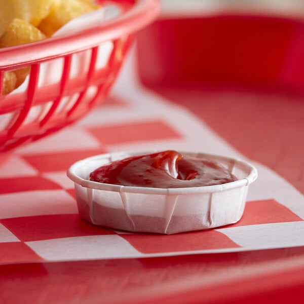 A small red Genpak container of ketchup with a basket of fries on a red and white checkered table.