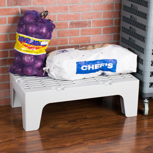 A white plastic Cambro dunnage rack with a bag of onions and a bag of potatoes on it.
