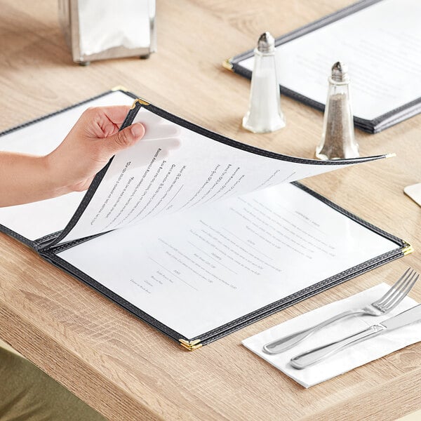 A person holding a Choice black four pocket menu cover with a table in the background.