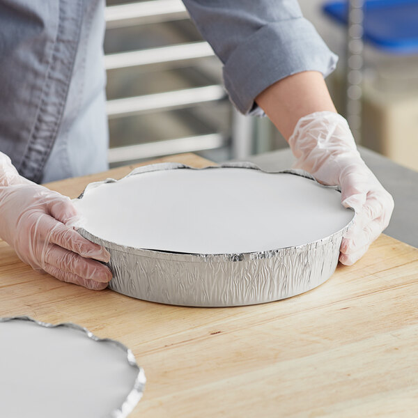A person in gloves holding a round white Choice foil take-out pan with white frosting.