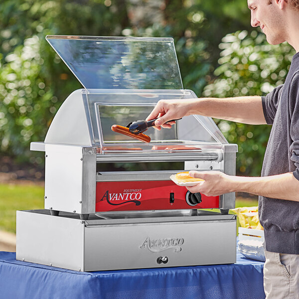 A man using an Avantco hot dog roller to cook hot dogs on a table in an outdoor catering setup.