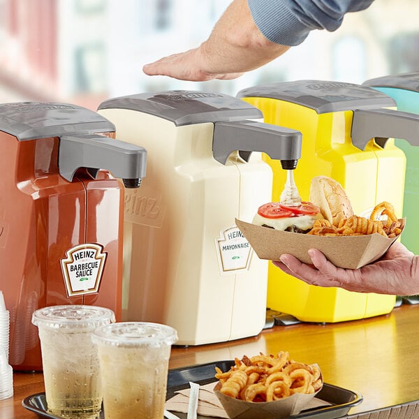 A person using a Heinz Keystone countertop mayonnaise pump to dispense sauce into a container of food.