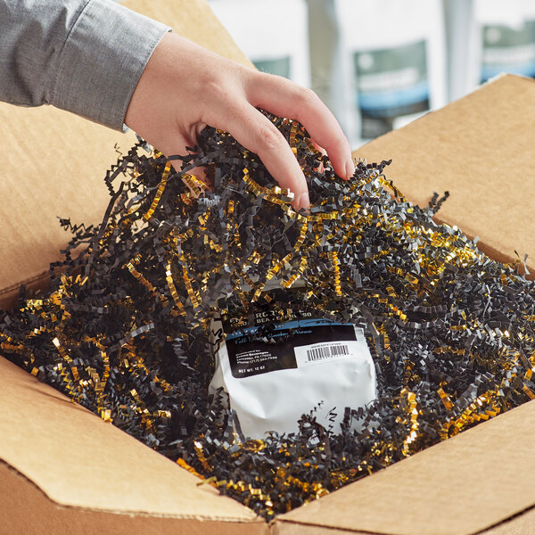 A hand holding a package with black and gold metallic crinkle cut paper in a cardboard box.