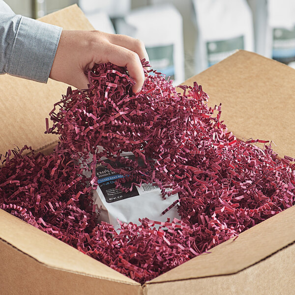 A hand holding a brown box filled with burgundy shredded paper.