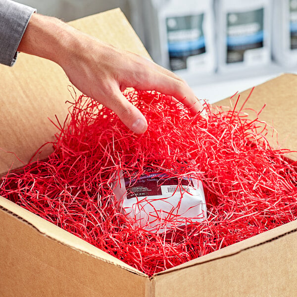 A hand putting a box with red Very Fine Spring-Fill paper shred into a cardboard box.