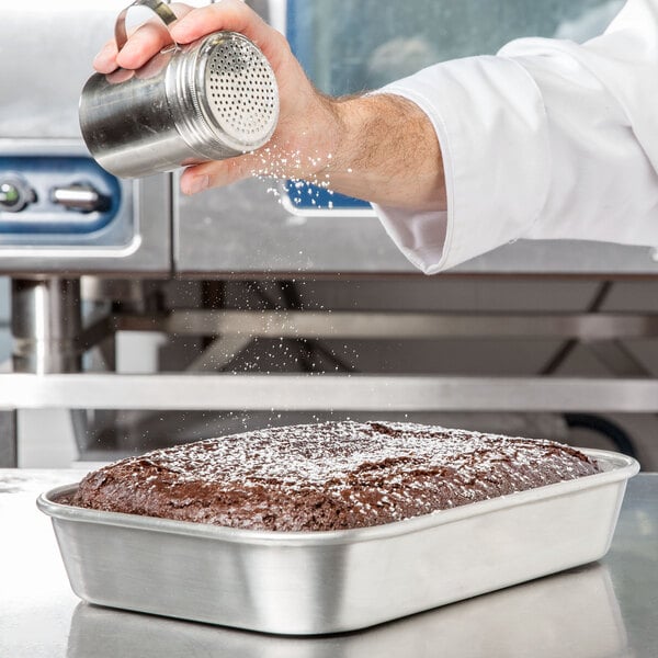 A person sprinkling powder onto a cake in a Vollrath aluminum baking and roasting pan.