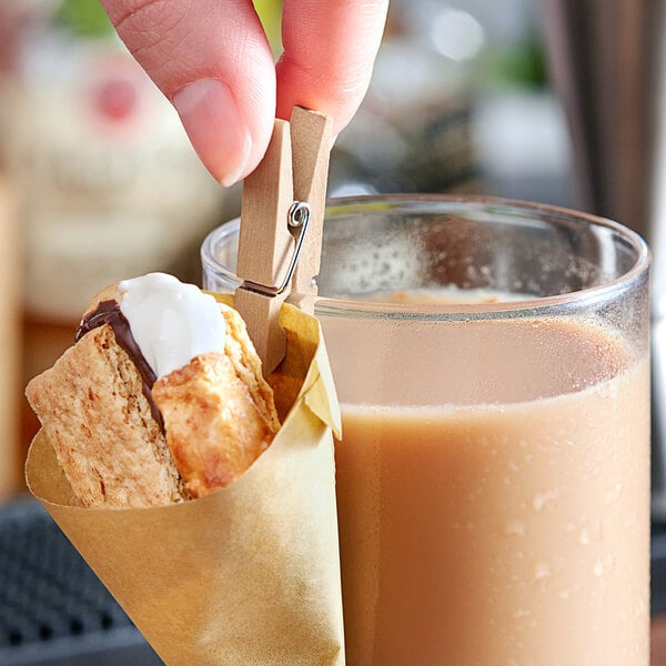 A hand using a mini wooden clothespin to attach a paper cone of food to a glass of liquid.