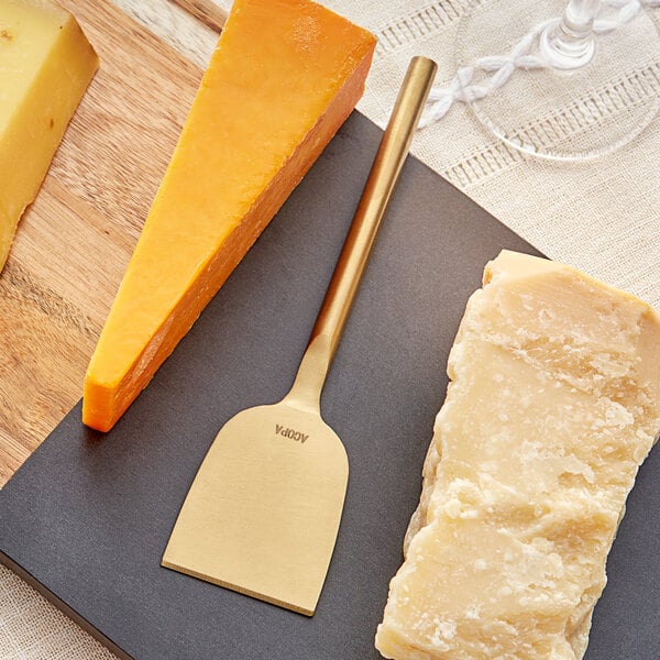 An Acopa gold stainless steel cheese knife cutting a piece of cheese on a cutting board.