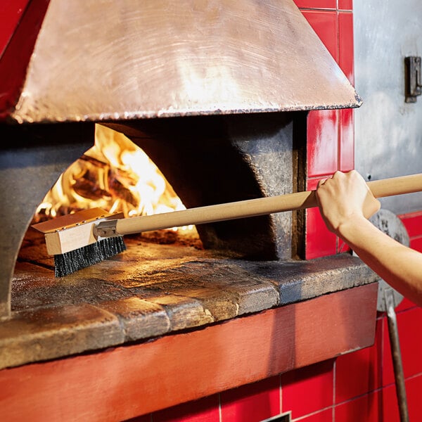 A person holding a Thunder Group pizza oven brush in a fire oven.