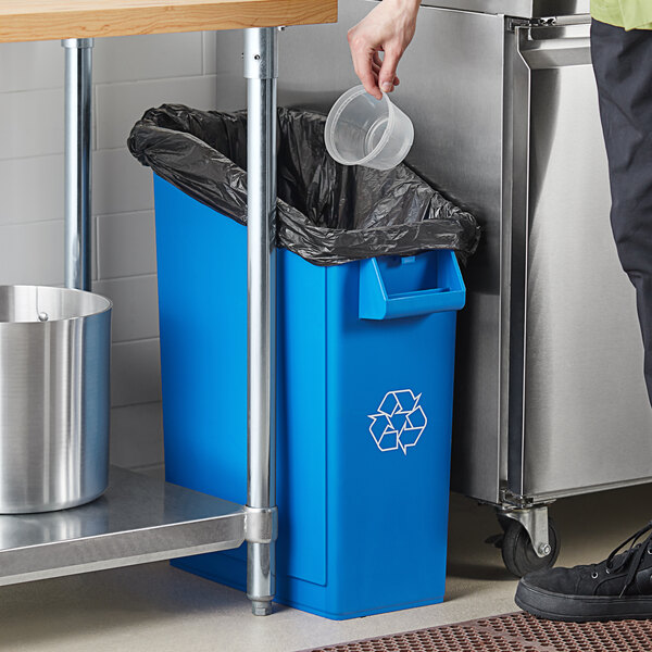 A person putting a plastic container into a blue rectangular recycling bin.