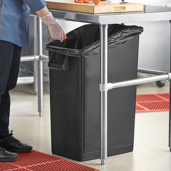 A person in a chef's outfit standing next to a Lavex black rectangular under-counter trash can.
