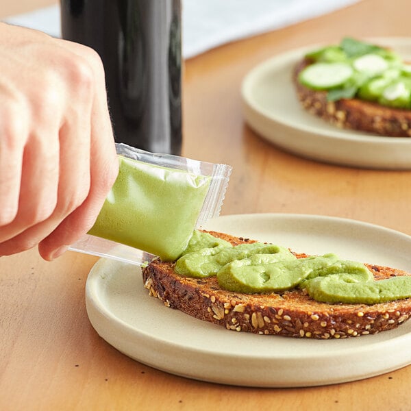 A hand using a Gator Pears avocado spread packet to put green sauce on a piece of bread.