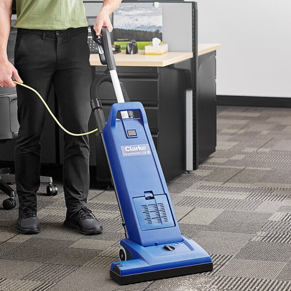 A person using a blue Clarke CarpetMaster 218 vacuum cleaner in a corporate office cafeteria.
